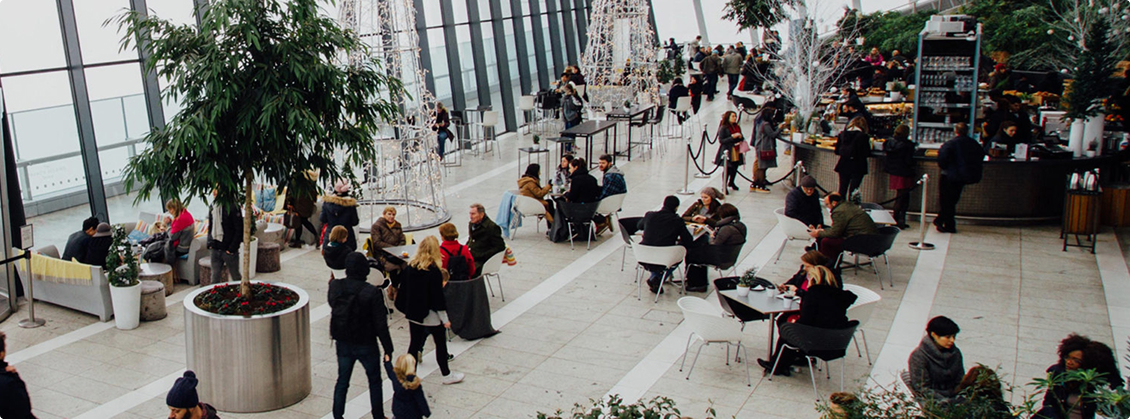 En ljus och modern inomhusmiljö med högt i tak och stora fönster. Människor sitter vid cafébord, promenerar eller står i kö vid ett café.